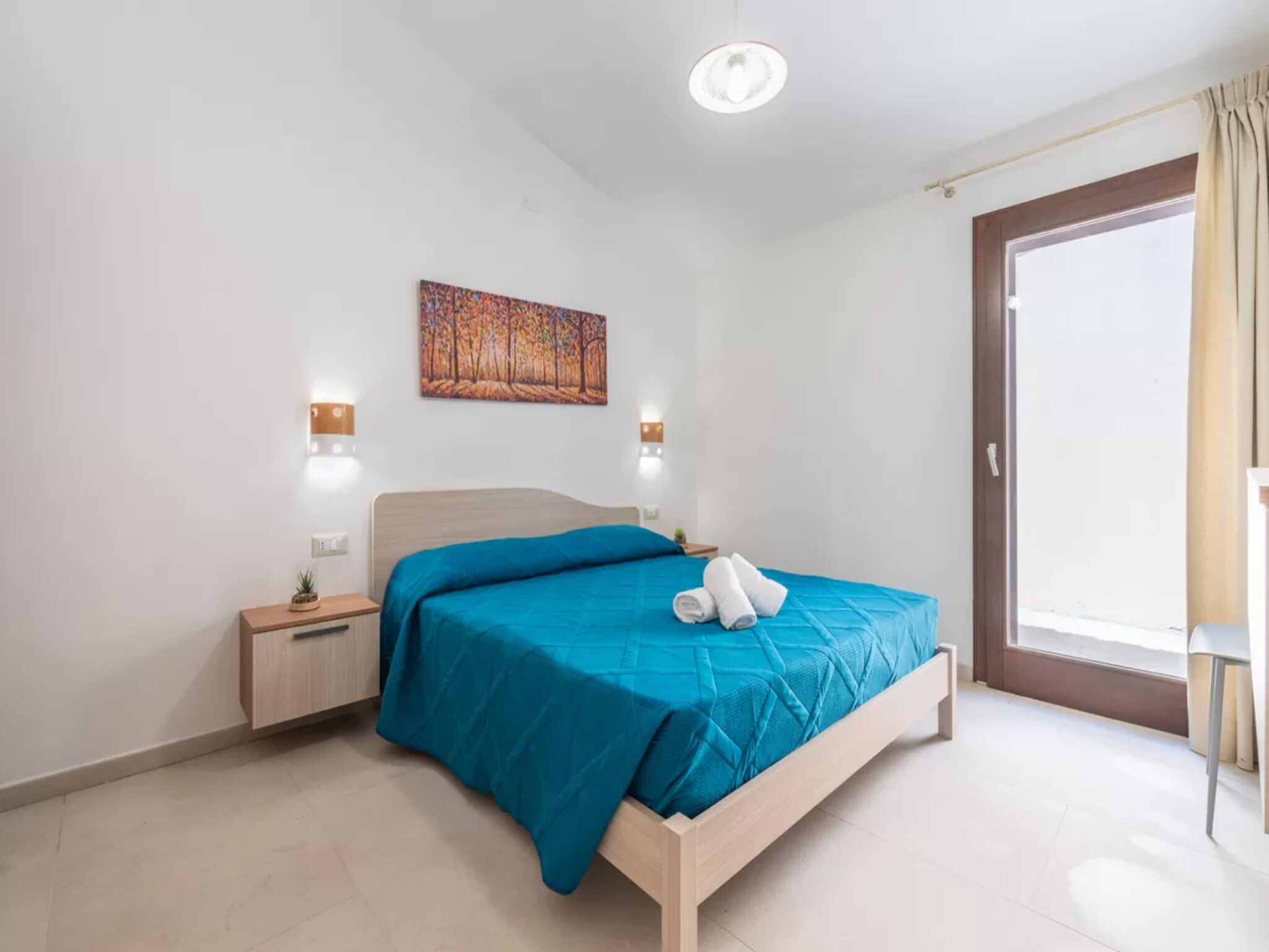 Un dormitorio con una cama, dos lámparas de noche, una mesita de noche, un cuadro en la pared y una ventana con cortinas.