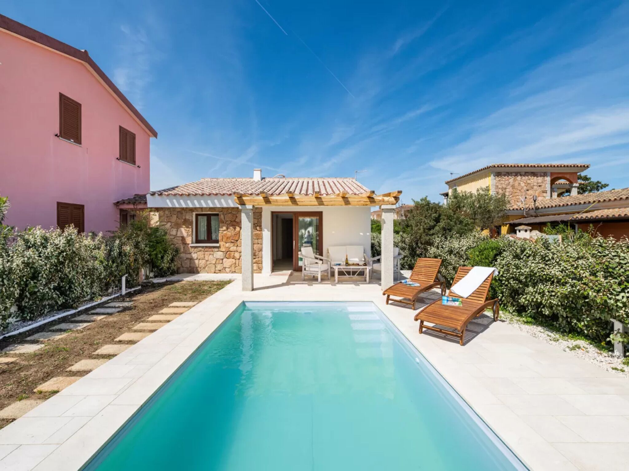Un espace aménagé au bord de la piscine, avec une piscine à l’eau d’un bleu limpide, un coin salon couvert avec des chaises, et un bâtiment en pierre surmonté d’un toit en tuiles.