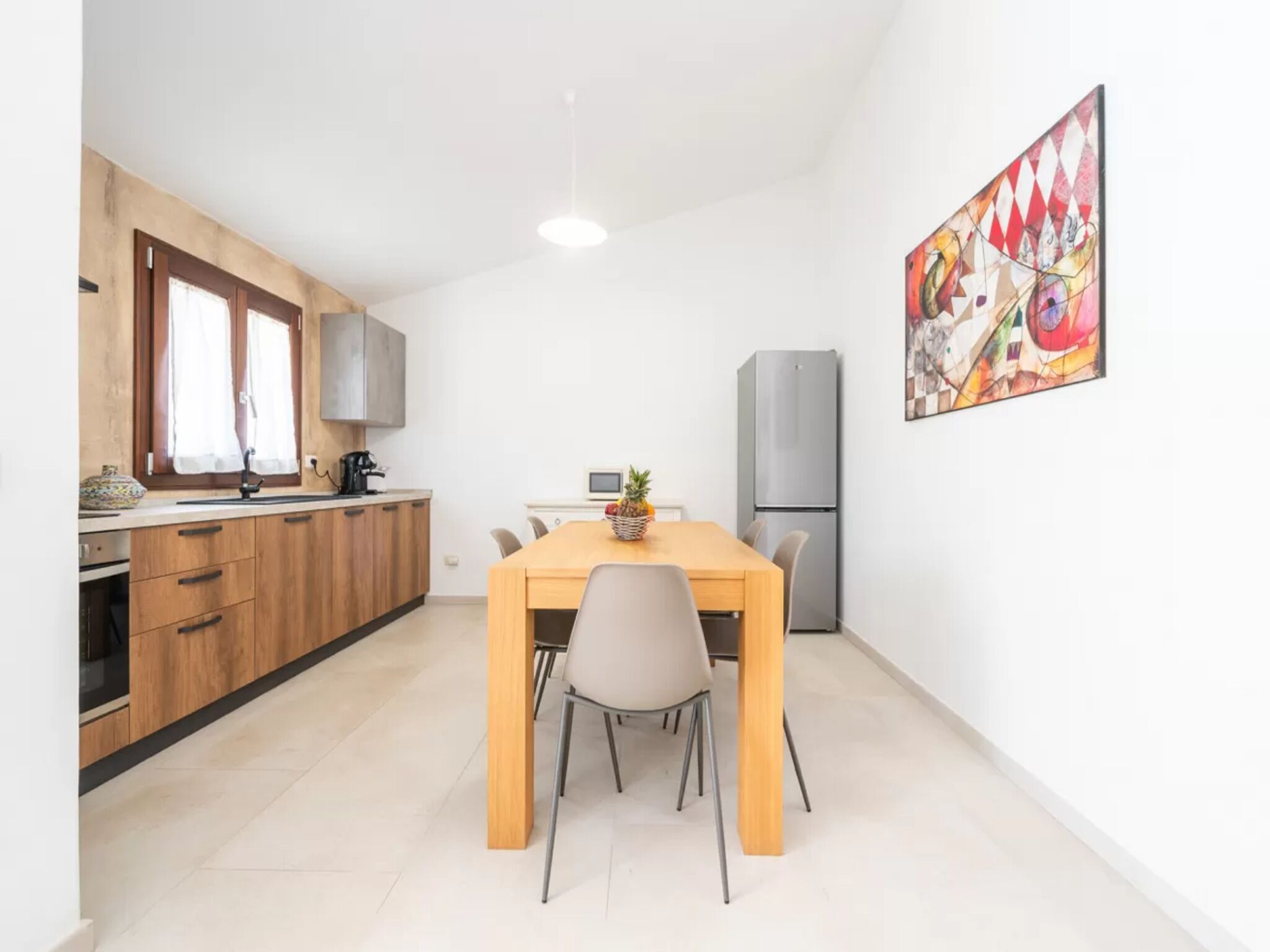 Een kleine keuken met houten kastjes, een eettafel met stoelen, een koelkast en een raam met gordijnen.