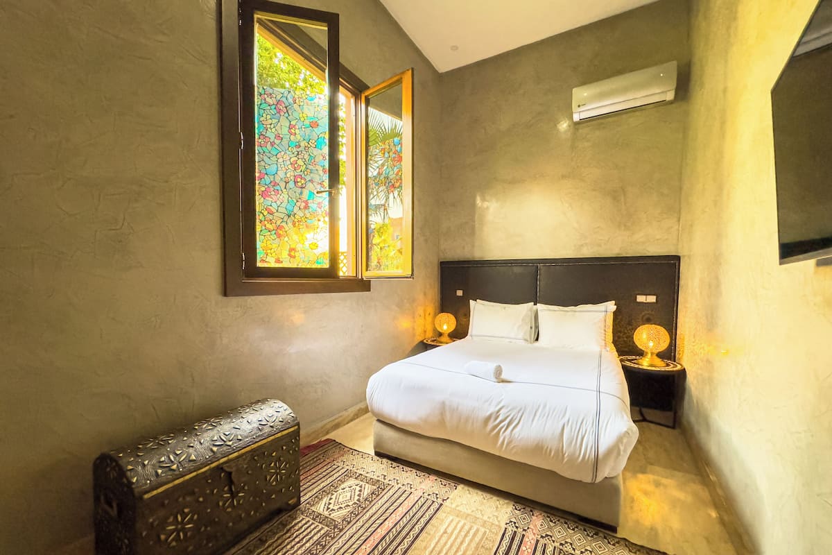 Une chambre à coucher comprenant un lit, une table de chevet, une commode décorative, un vitrail et un appareil de climatisation.