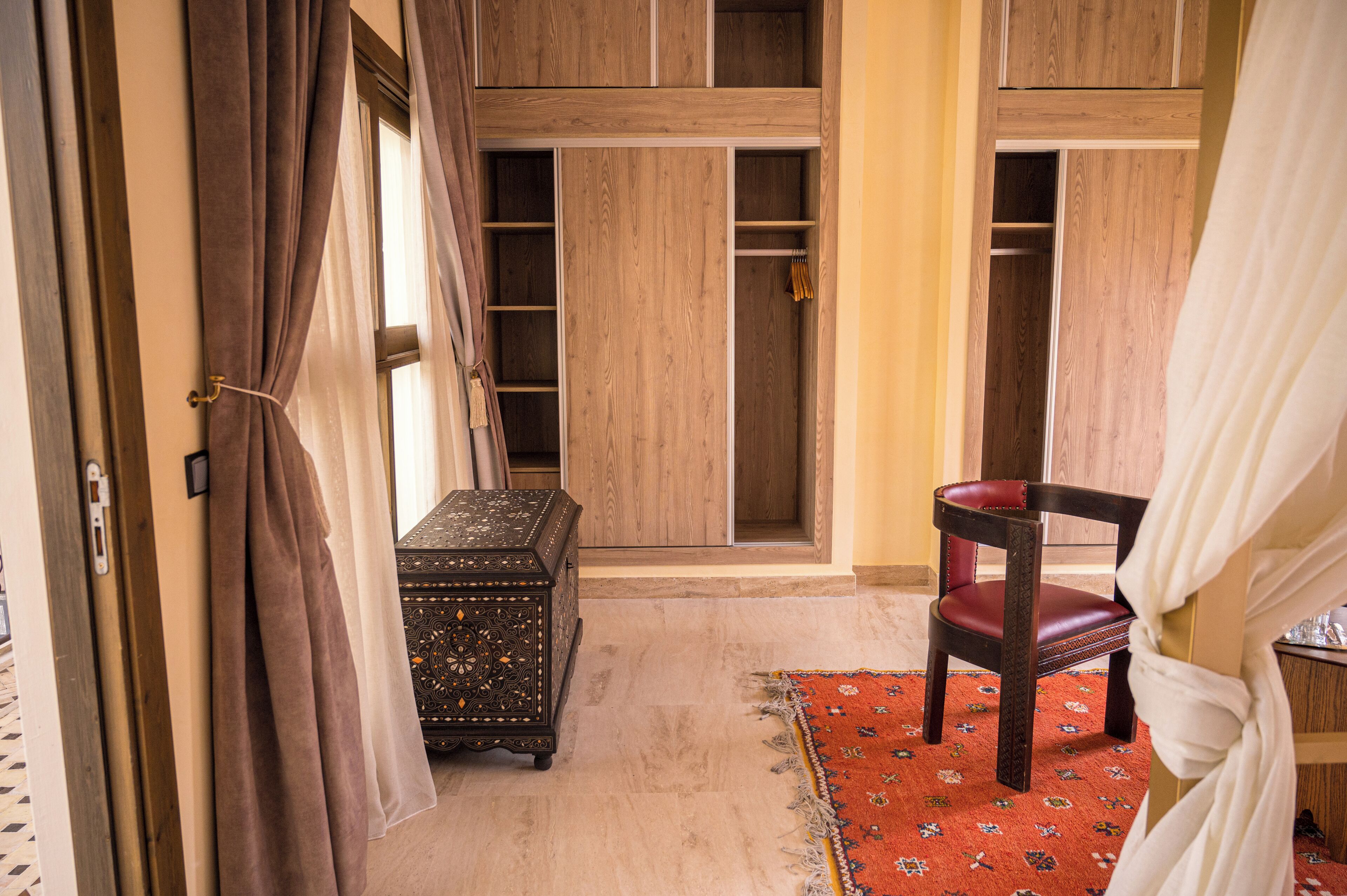 Una habitación con un armario de madera, un tapete rojo estampado, un cofre de madera y una silla con cojín rojo.
