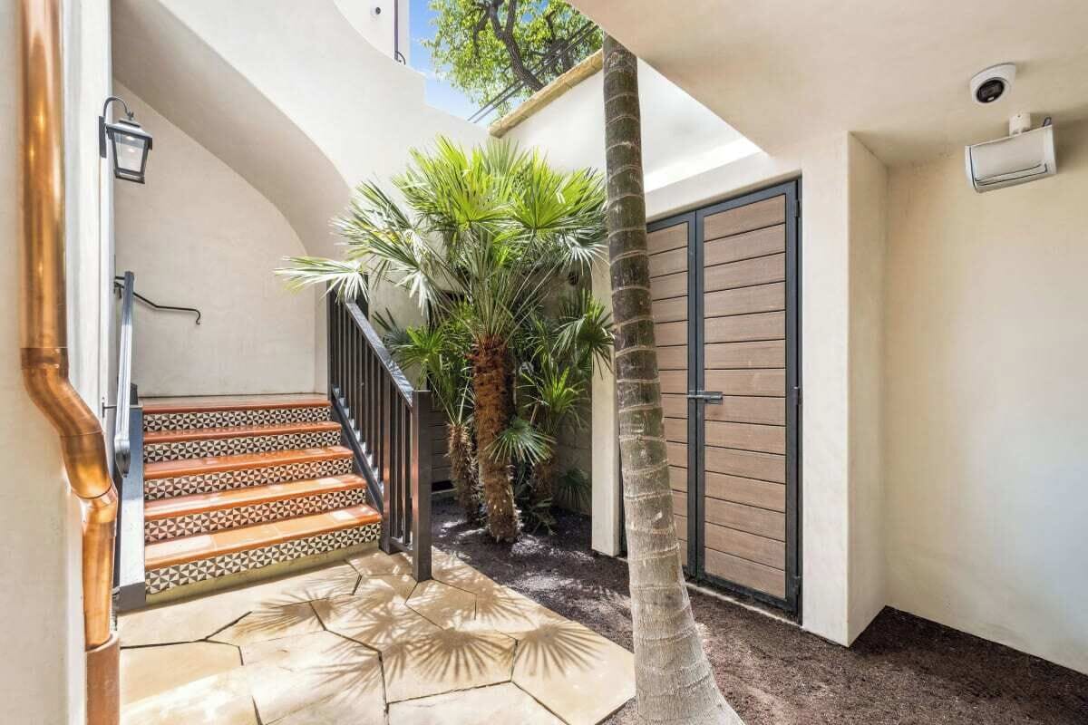 Eine Treppe mit gemusterten Fliesen führt zu einer Tür, umgeben von Palmen und einer gefliesten Terrasse.
