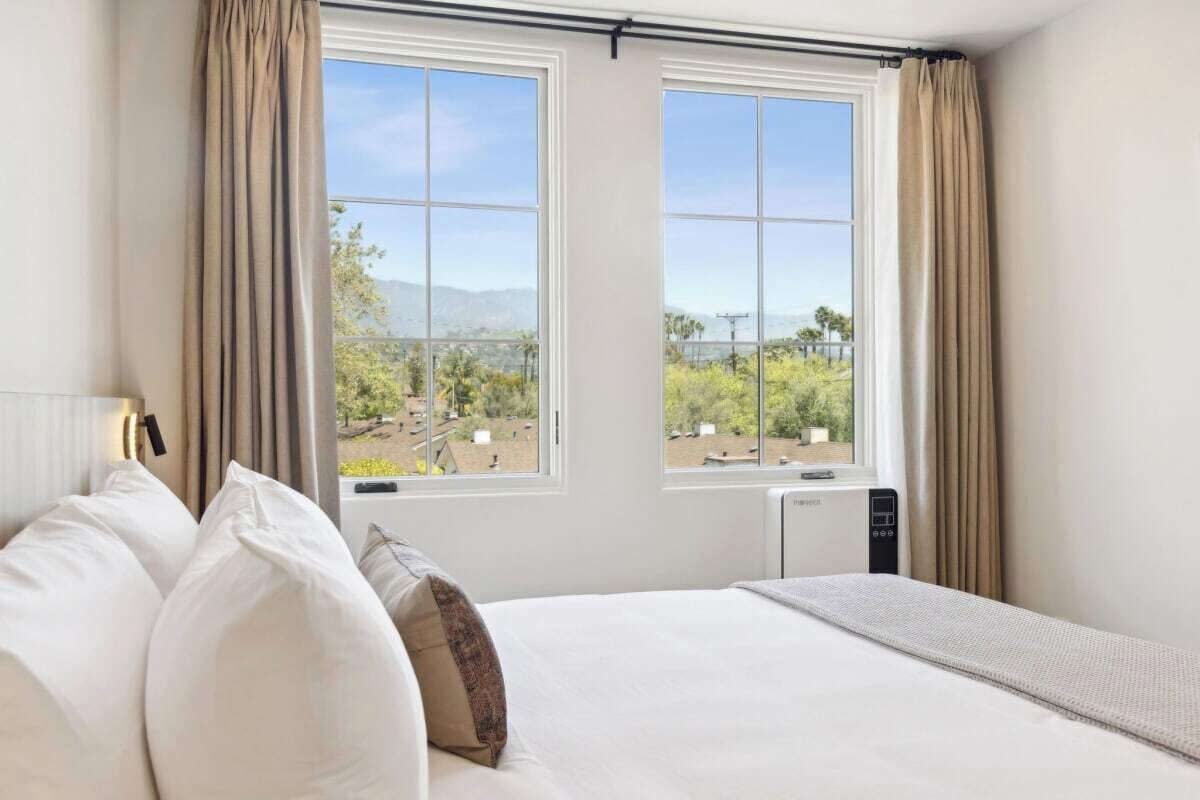 Ein Hotelzimmer mit einem großen Bett, zwei Fenstern mit Vorhängen und Blick auf eine Berglandschaft.