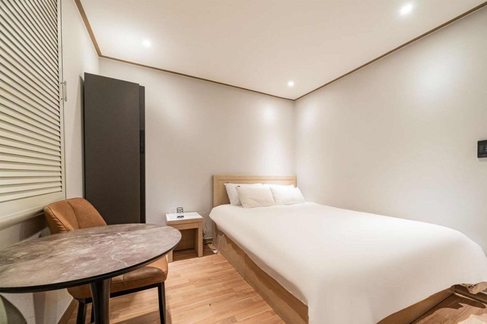 Habitación de hotel con cama, una mesita, una silla, una mesita de noche y una lámpara fijada a la pared.