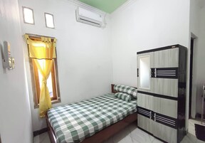 Quarto pequeno com uma cama de solteiro, colcha xadrez, janela com cortinas amarelas, guarda-roupa com espelho e ar condicionado na parede.