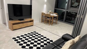 Un salon moderne avec un canapé en cuir noir, un meuble de rangement en bois intégrant un téléviseur, un coin repas avec une table et des chaises, et un tapis à motifs.