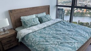 Une chambre à coucher avec un grand lit, une table de chevet avec une lampe et une vue sur le paysage urbain par la fenêtre.