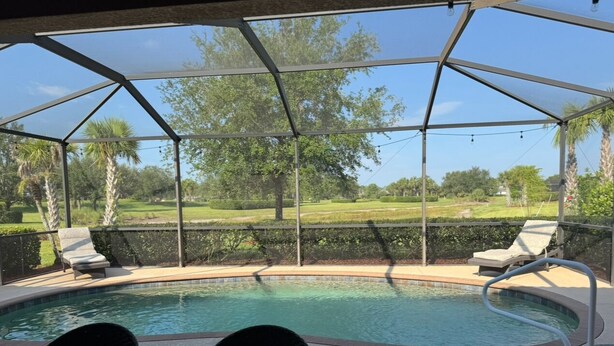 Piscina exterior coberta com espreguiçadeiras e uma mesa, rodeada por uma vedação metálica e árvores.