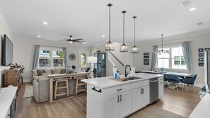 A modern kitchen with a central island, a dining area with blue chairs, and a living room with a sofa and wooden furniture. - 4BR Home with Deck & Grill Fun + Free Attraction Tickets! (Santa Rosa Beach)