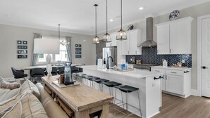A modern kitchen with a central island, bar stools, and a dining area with a wooden table and chairs. - 4BR Home with Deck & Grill Fun + Free Attraction Tickets! (Santa Rosa Beach)