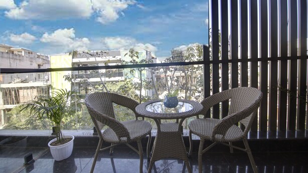 Un balcón con sillas de mimbre y una mesa, una planta en maceta y vistas a edificios y árboles.