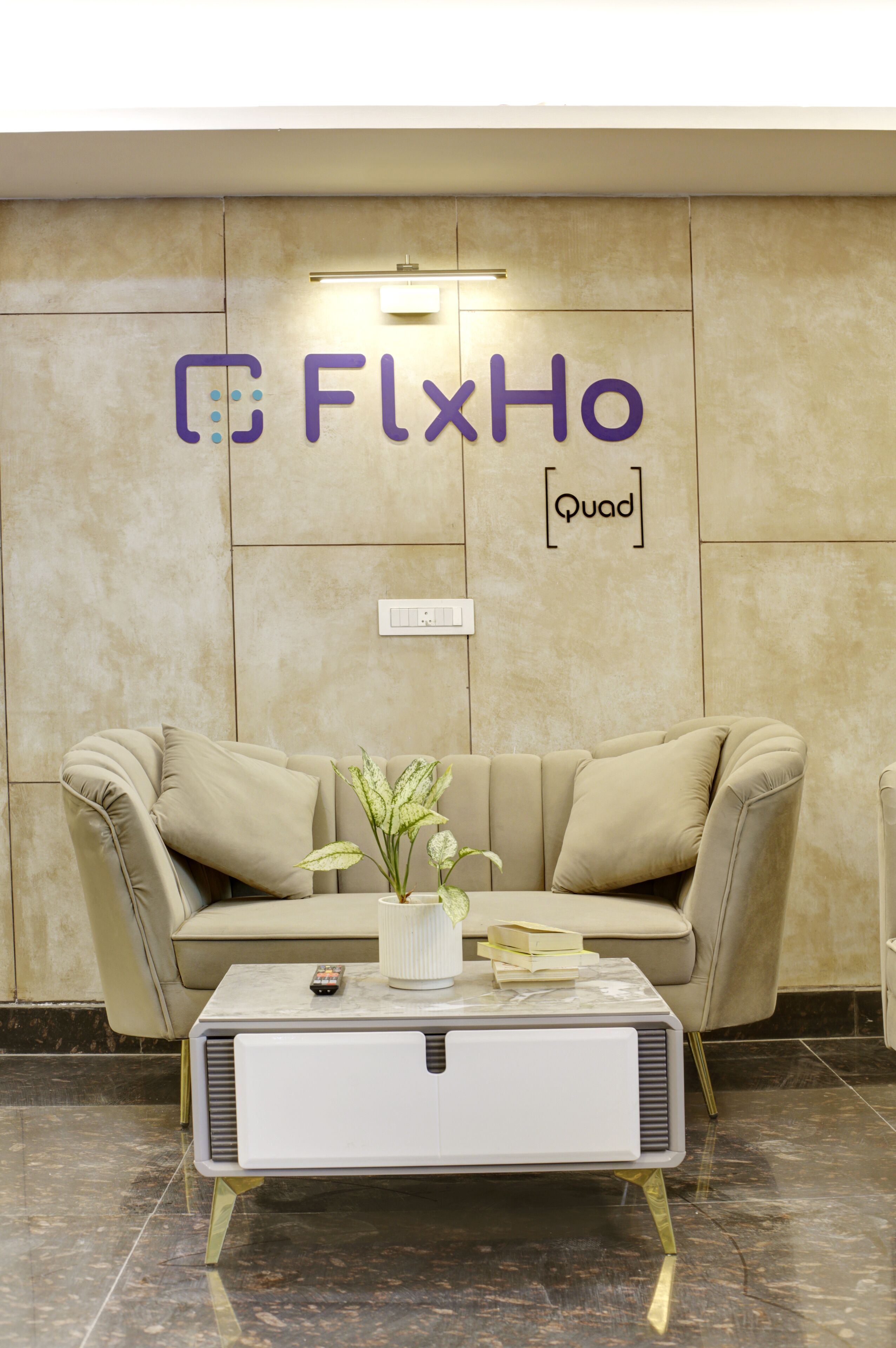 Un hall d’entrée moderne avec un canapé beige, une table basse blanche et une enseigne fixée au mur portant l’inscription « FlexHo Quad ».