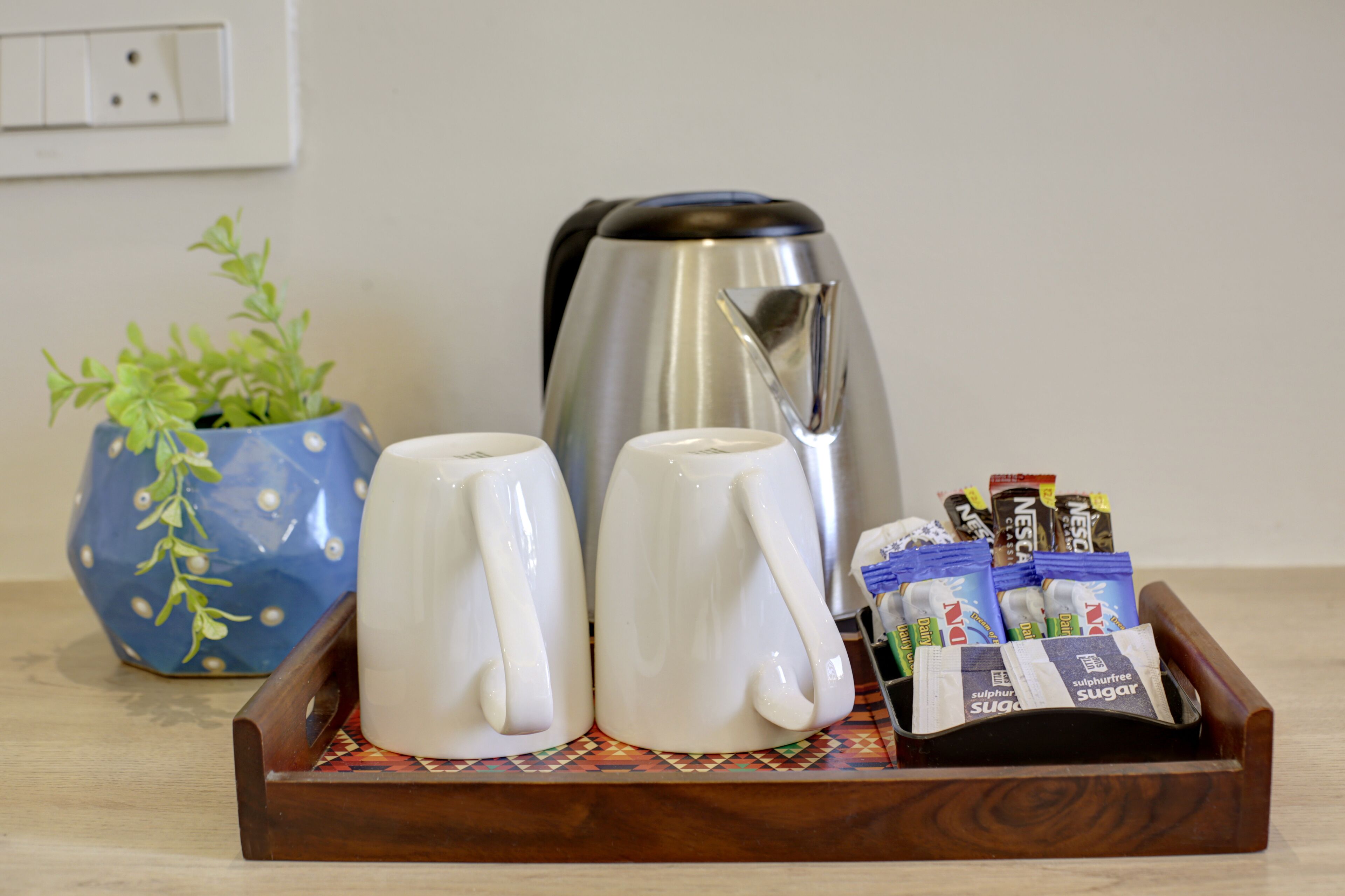 A tray with two white mugs, a stainless steel kettle, a blue decorative pot with green plants, and several packets of sugar and coffee creamer.