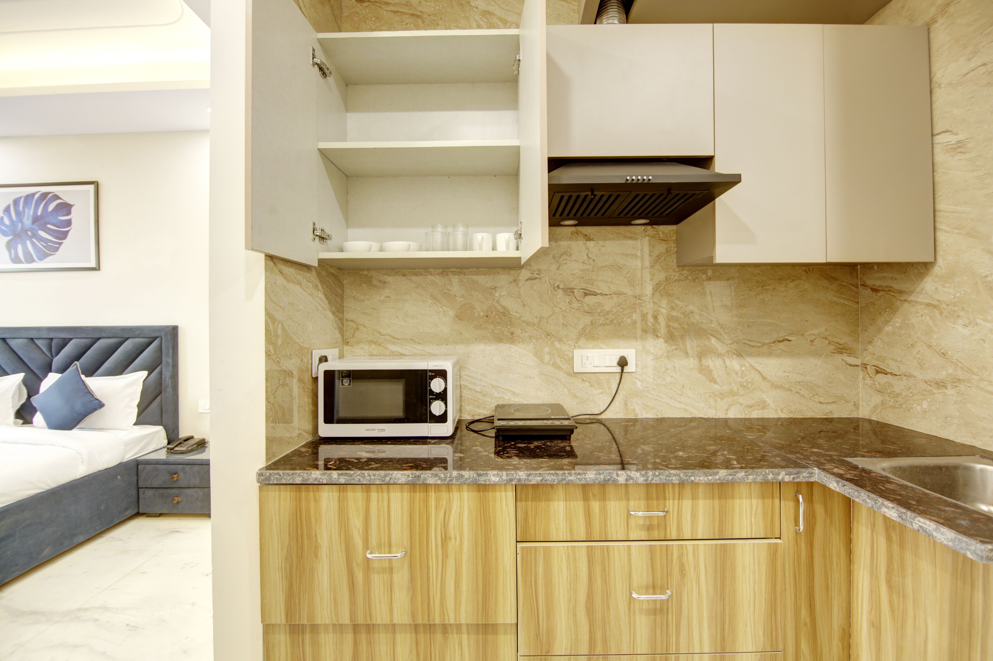 A compact kitchen with a microwave, a toaster, and a sink. There is a bed with a headboard and a nightstand with a phone. The kitchen has wooden cabinets and a marble backsplash.