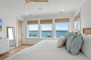 A bedroom with a large bed, a dresser, a mirror, and a view of a body of water through large windows.