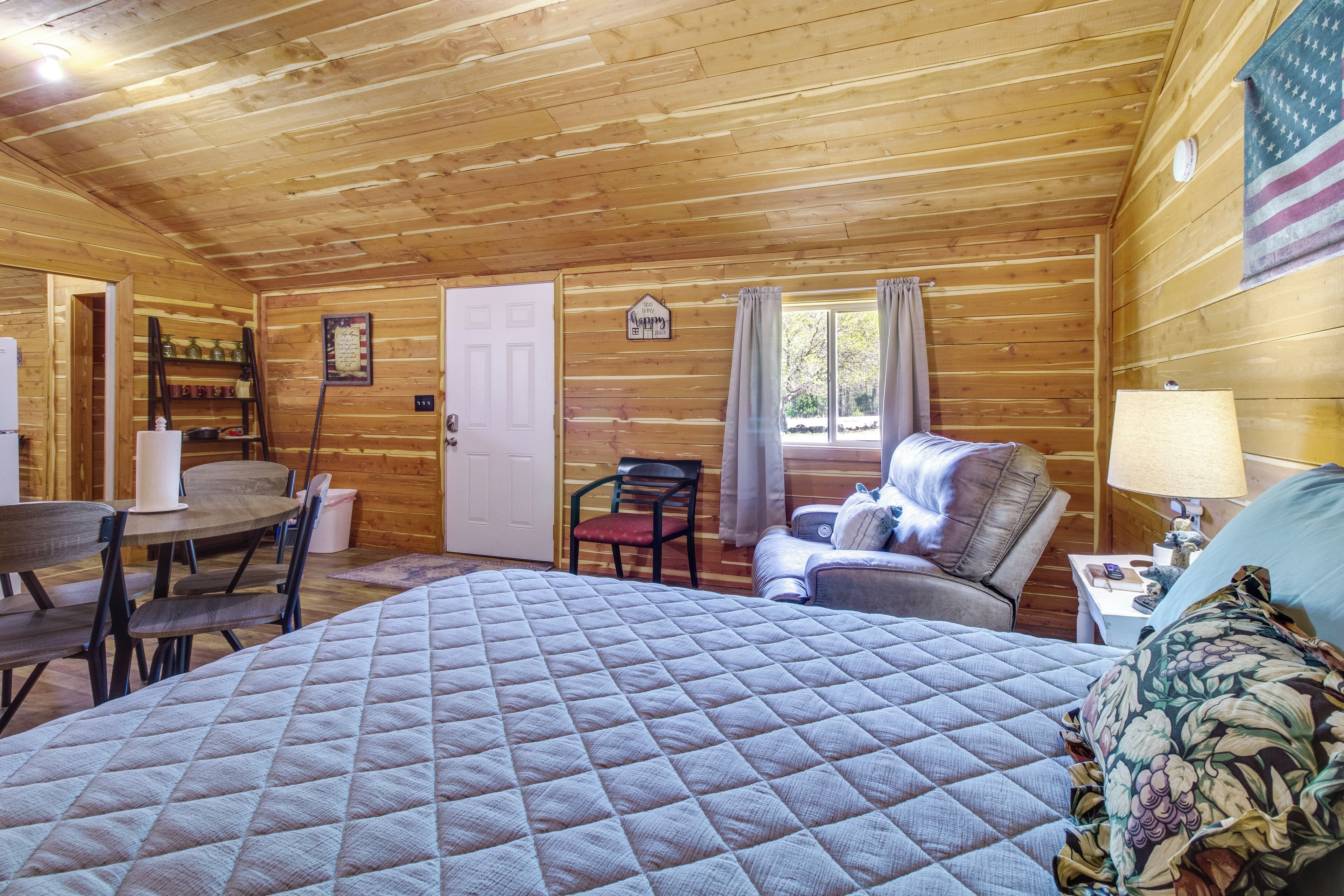 A cozy cabin room with a large bed, a dining table, chairs, a small table with a lamp, and a window with curtains.