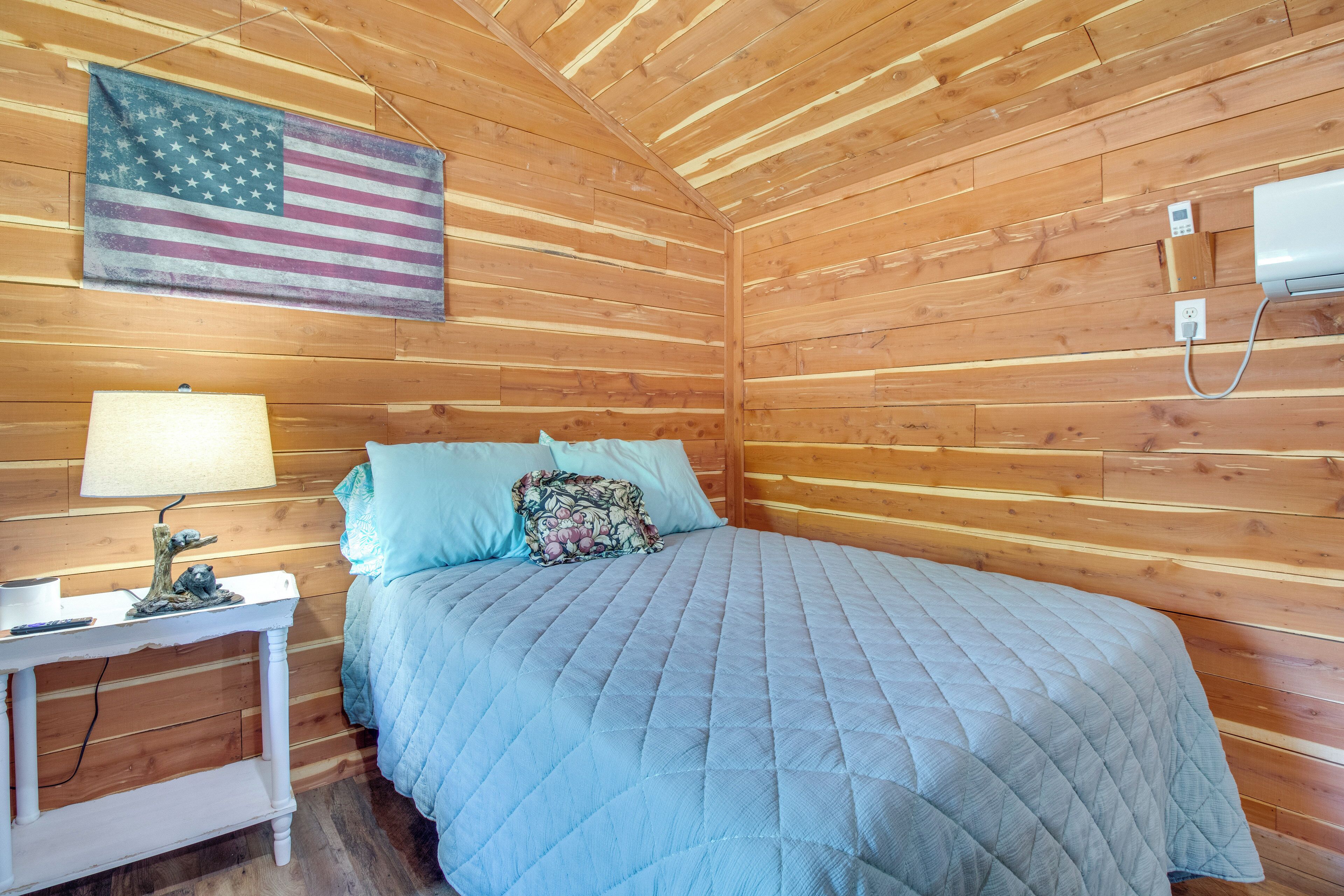 A wooden cabin room with a bed, a nightstand, a lamp, an American flag, and a thermostat.