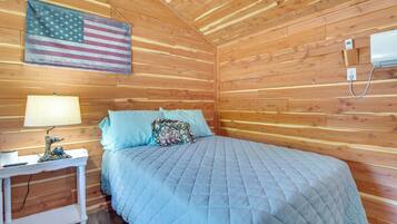 A wooden cabin room with a bed, a nightstand, a lamp, an American flag, and a thermostat.