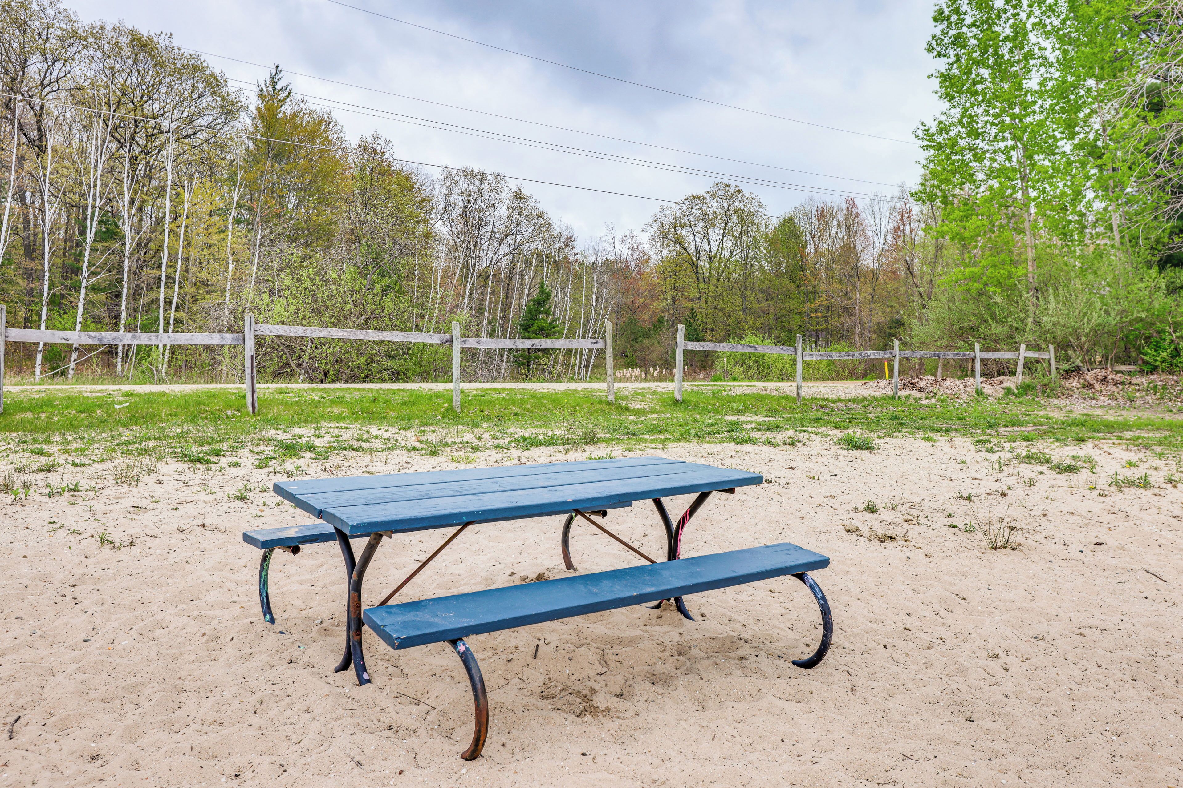 A picnic table in a sandy area with a wooden fence and trees in the background.