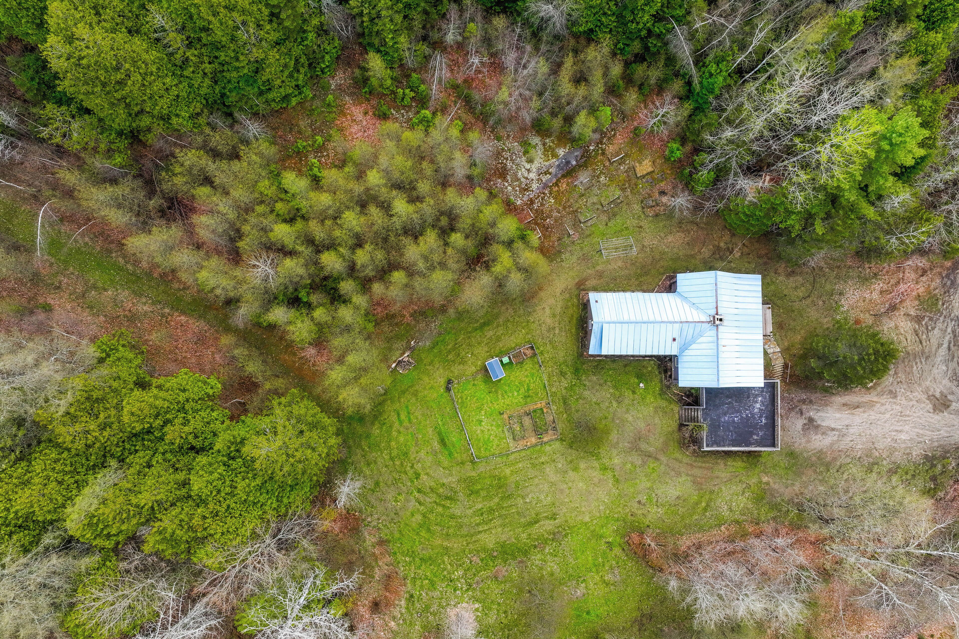 An aerial view of a house surrounded by dense forest, with a clear area in front and a blue-roofed structure.
