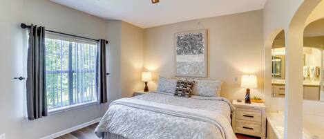 A bedroom with a large bed, a ceiling fan, a window with curtains, a bedside table with a lamp, and a bathroom with a mirror and sink.