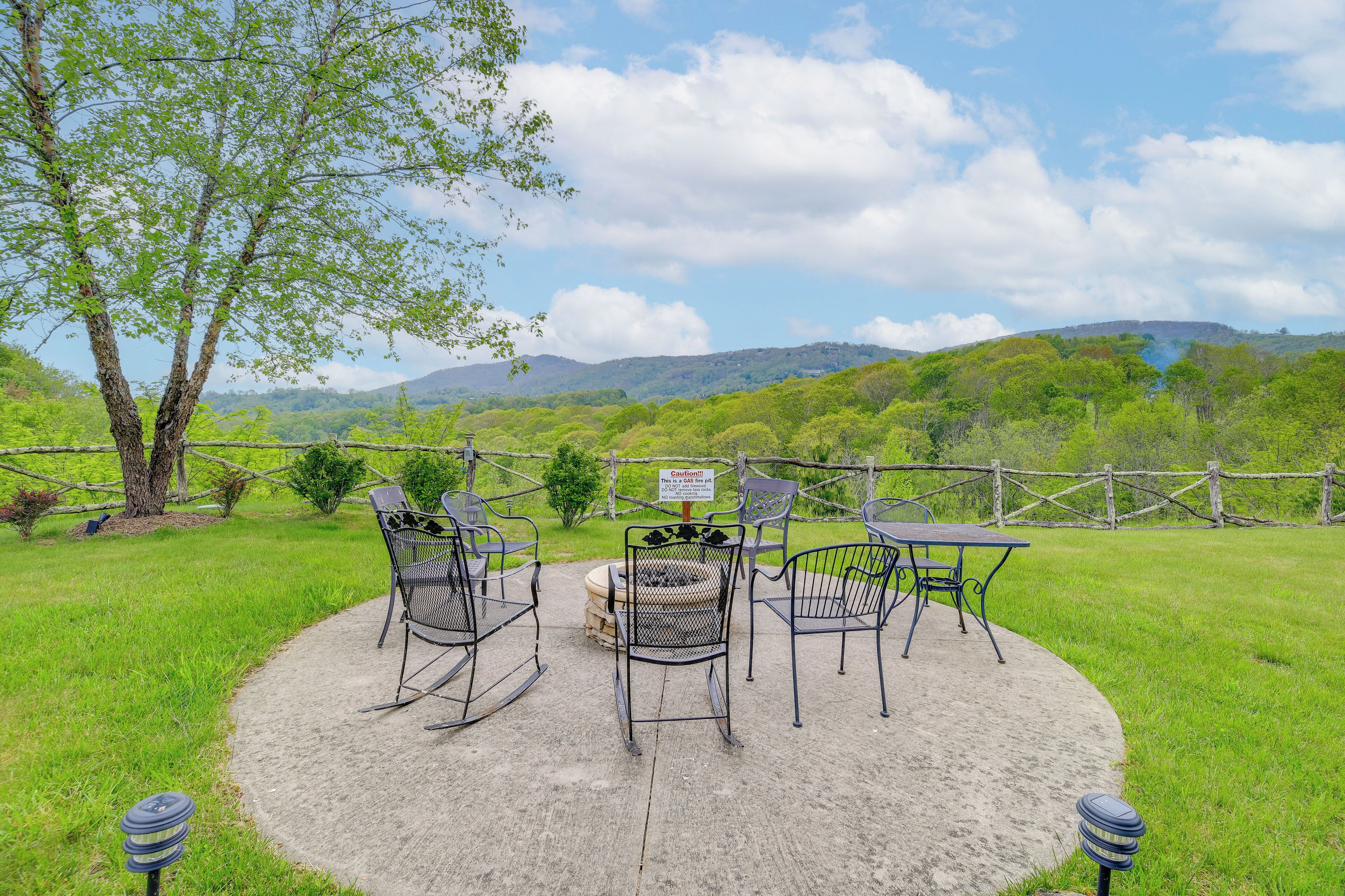 A patio with metal chairs and a table, surrounded by a wooden fence, overlooking a green landscape with hills.