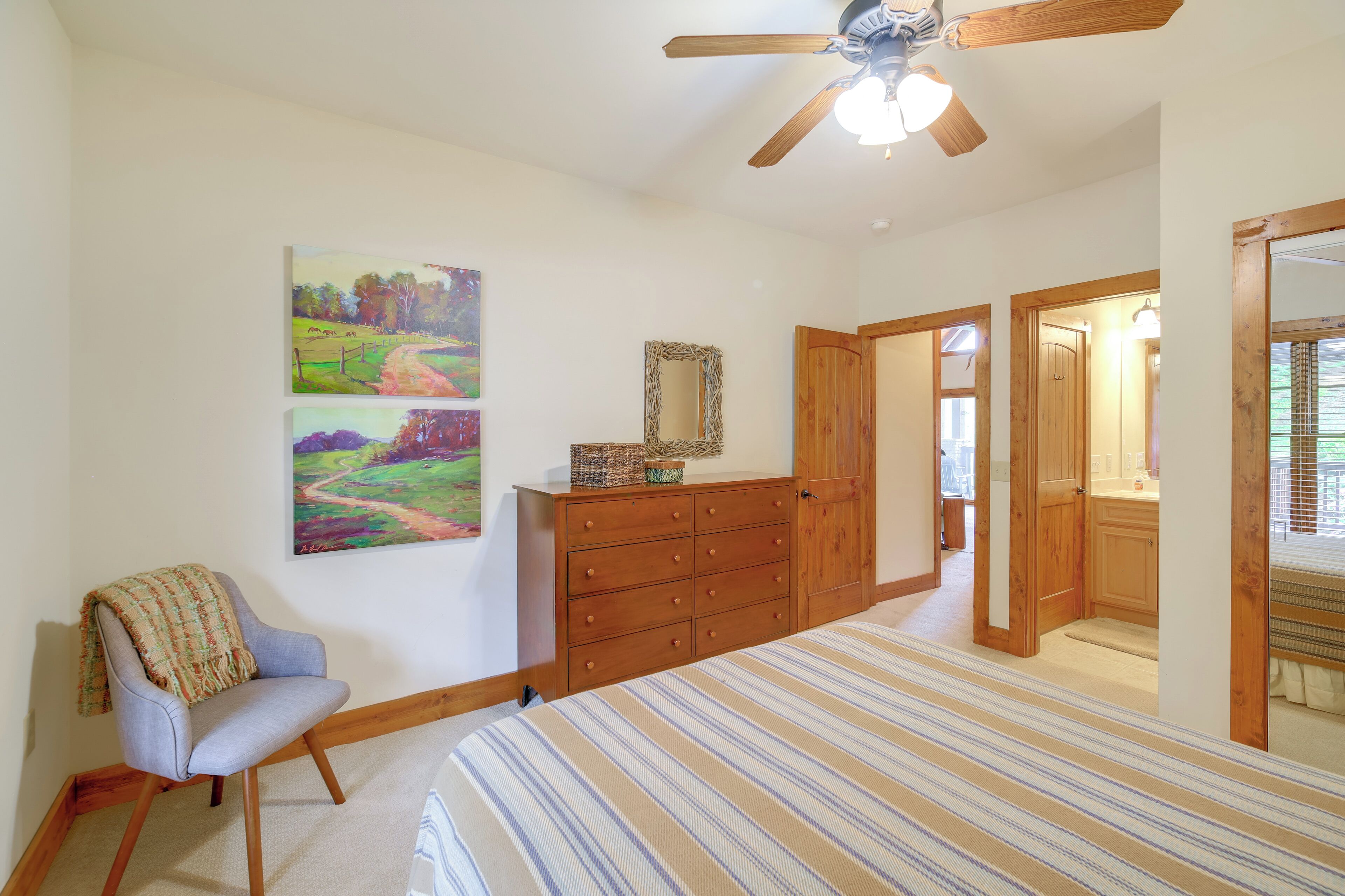 A bedroom with a bed, a chair, a dresser, a ceiling fan, and two paintings on the wall.