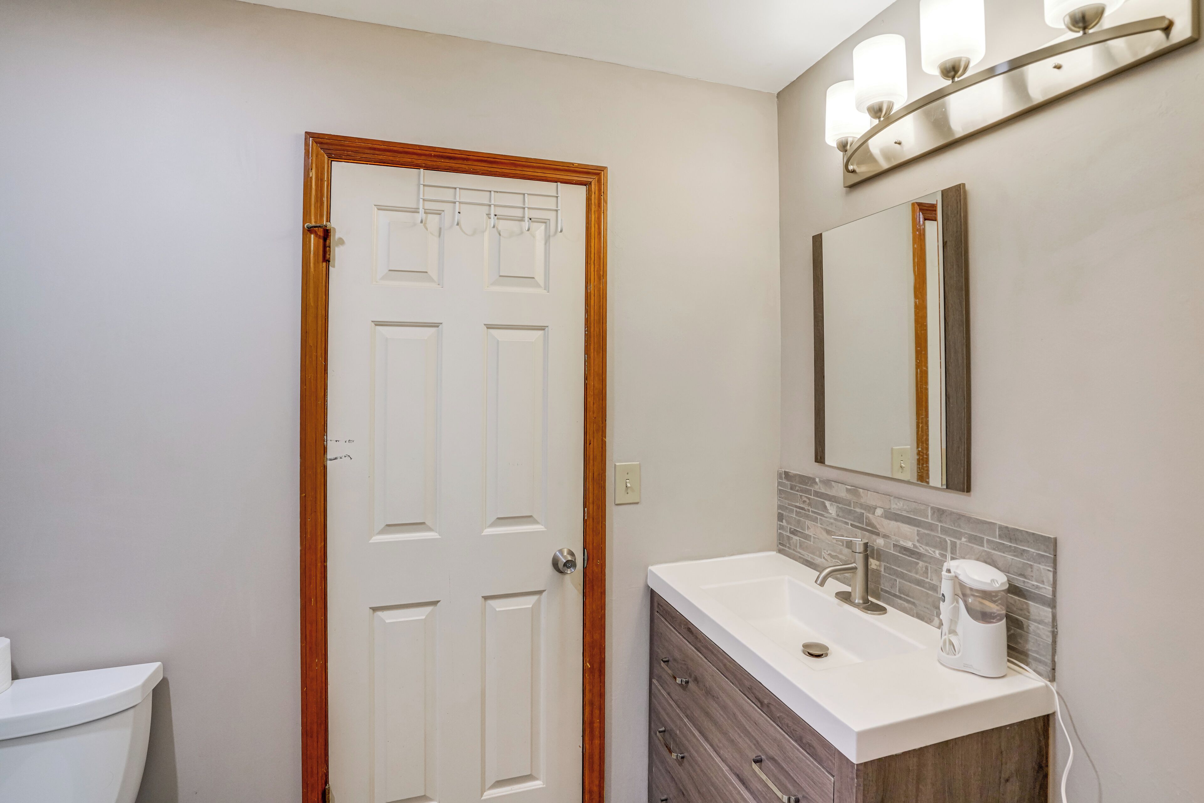 Un baño con inodoro blanco, puerta de madera, lavamanos blanco con grifo, espejo y dispensador de toallas.