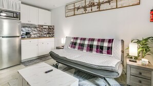 Un espace de vie compact comprenant un coin cuisine, un lit avec une couverture matelassée, une table de chevet avec une lampe et une œuvre d’art accrochée au mur.