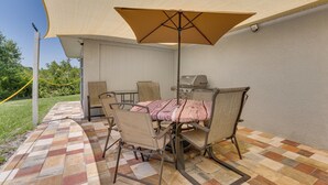 Une terrasse avec une table et des chaises sous un grand parasol, un barbecue et un sol à carreaux.