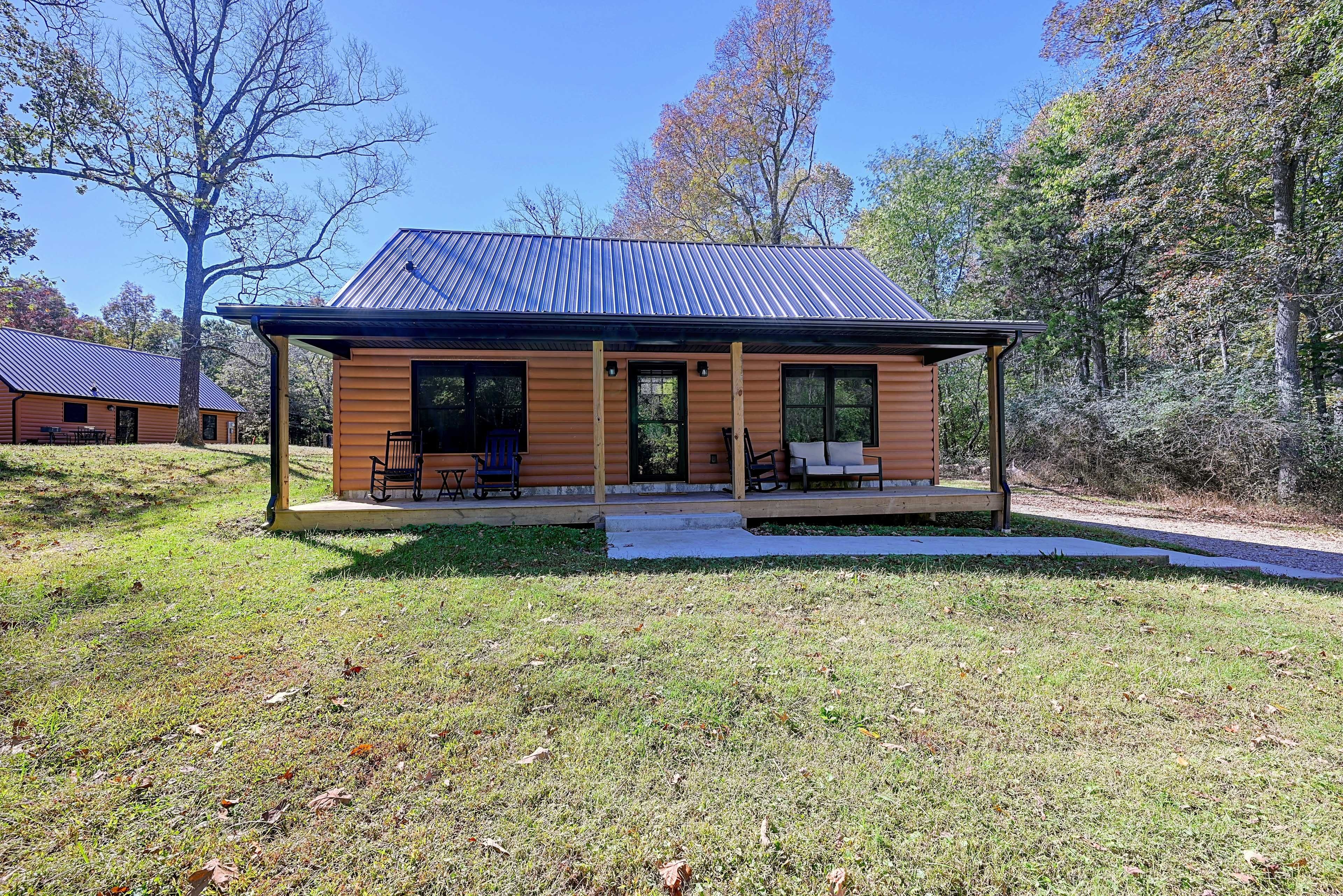 A single-story log cabin with a covered porch, a small deck, and a clear view of the surrounding trees.