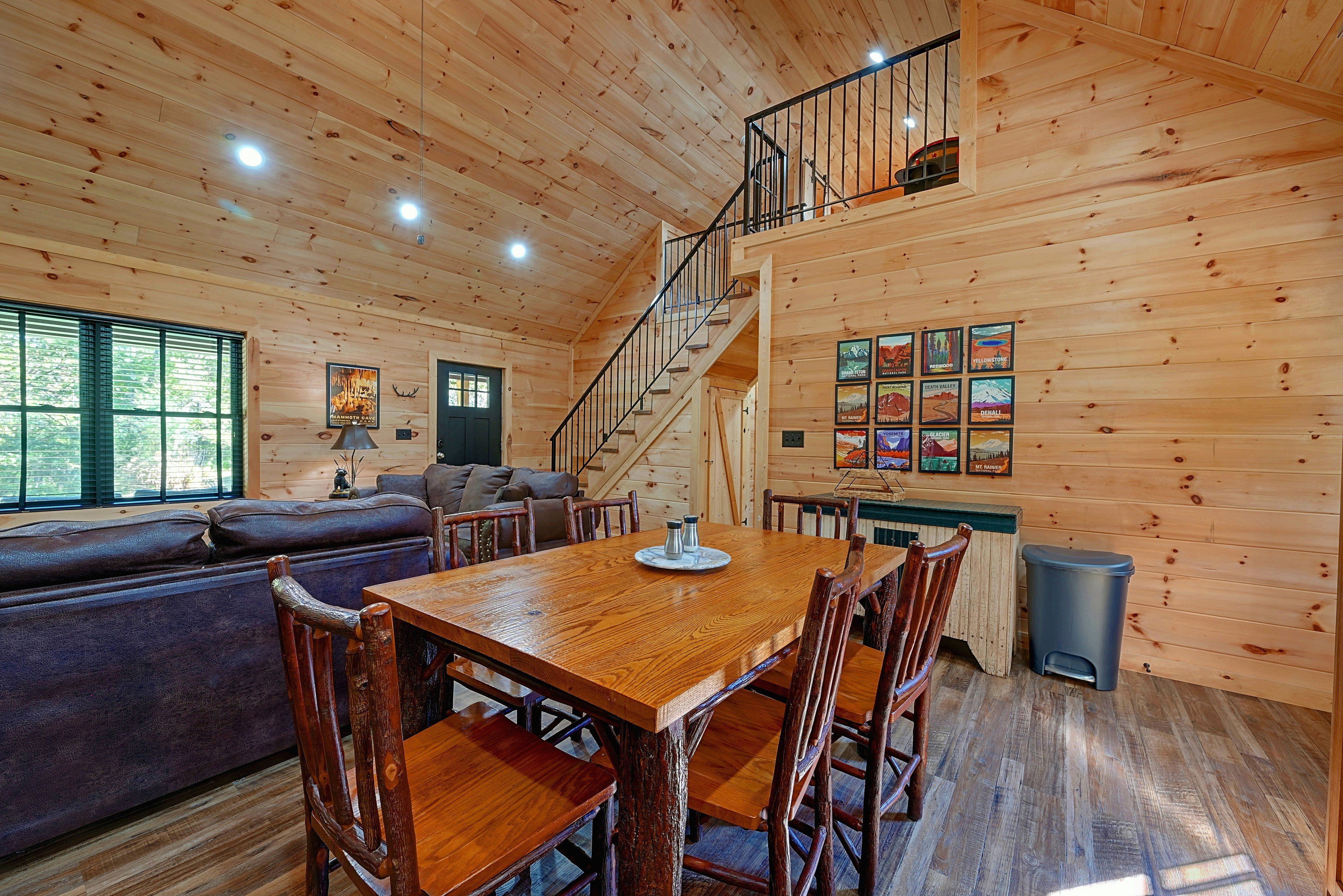A cabin interior with a wooden dining table, chairs, a sofa, a staircase, and a wall with framed pictures.