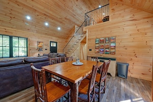 Interior de una cabaña con una mesa de comedor de madera, sillas, un sofá, una escalera y un muro con cuadros enmarcados.