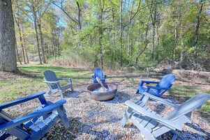 Un camping con sillas Adirondack, un hogar para fuego y un entorno boscoso natural.