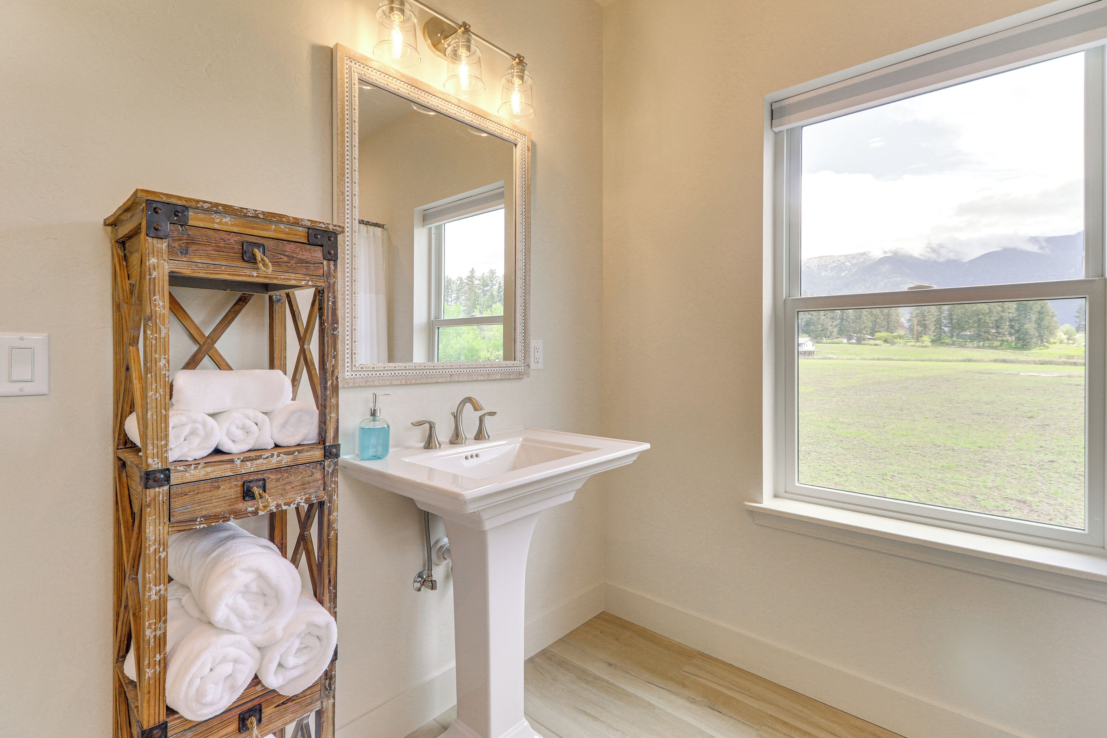 A bathroom with a wooden towel rack, a white sink, and a large mirror with lights.