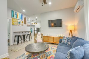 A modern living room with a blue sofa, a round coffee table, and a colorful rug. There are bar stools, a wicker chair, and a TV mounted on the wall.