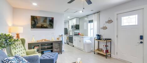 Una sala de estar moderna con un sofá, un reposapiés y una mesa de centro, una cocina con armarios y electrodomésticos blancos, y un ventilador de techo.