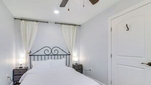 A neatly made bed with white linens, two bedside tables with lamps, a ceiling fan, and a white door with a handle.