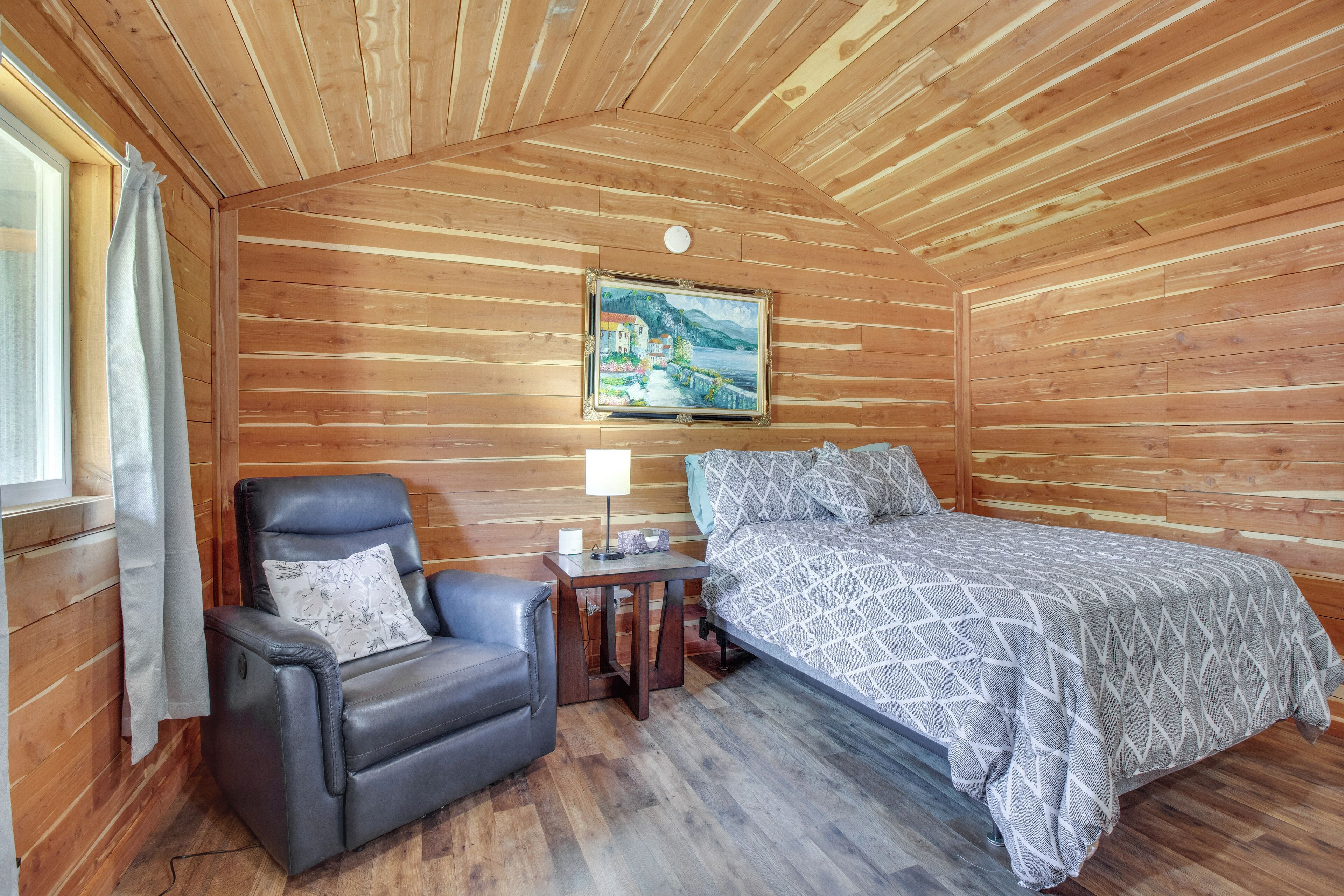 A wooden cabin room with a bed, a leather chair, a nightstand, and a painting on the wall.