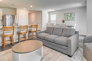 Uma sala de estar moderna com um sofá cinza, cadeiras de madeira, uma mesa de centro redonda e uma área de jantar com uma mesa e cadeiras brancas.