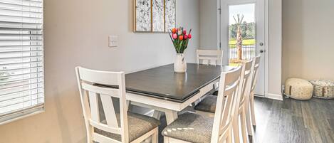 A dining area with a dark wooden table and white chairs, a vase with red flowers, and a door leading outside.
