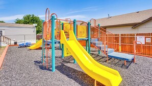 A playground with slides and climbing structures, surrounded by a wooden fence and a building in the background. - 13 Mi to Okc: Edmond Home w/ Pool Access! (Edmond)