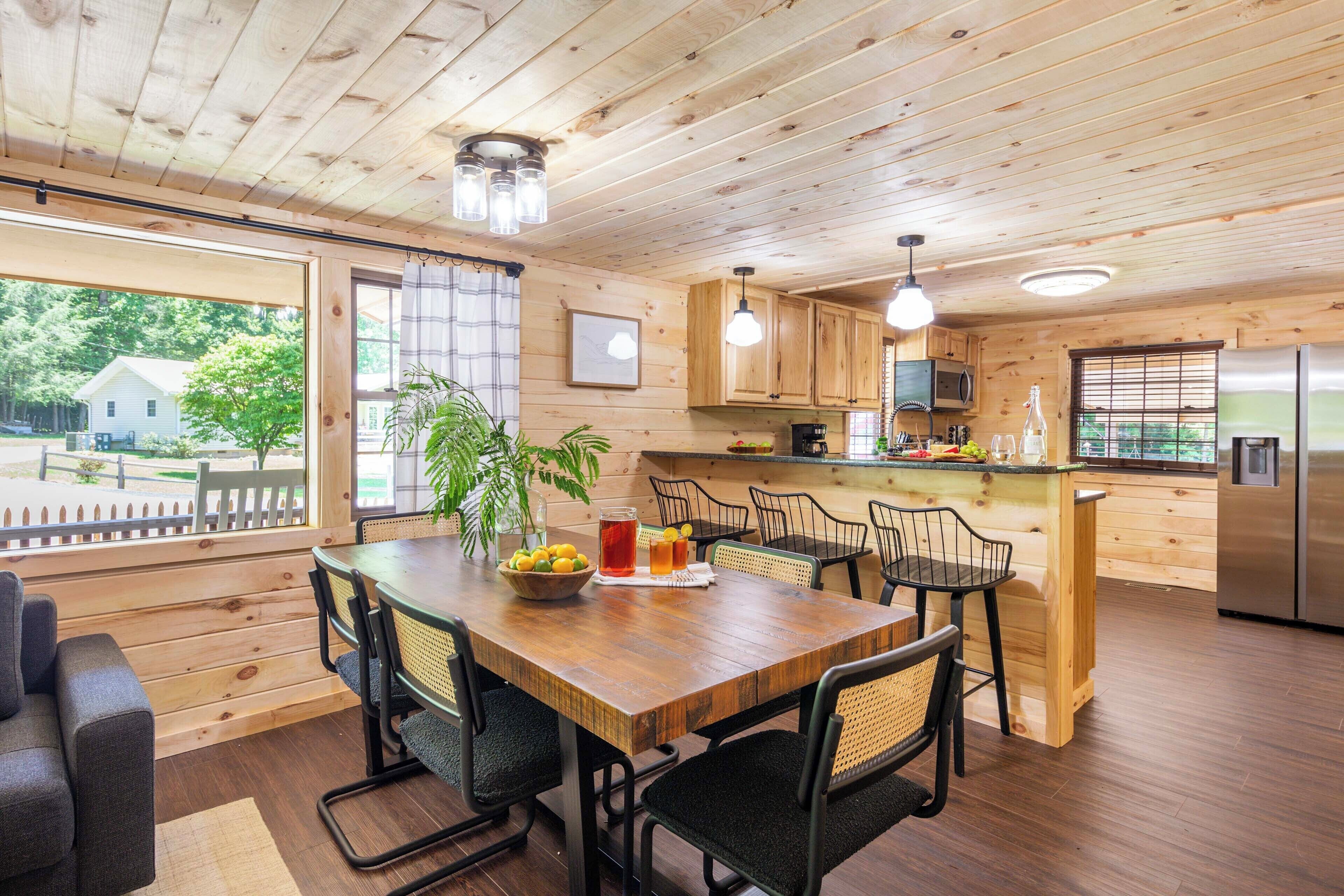 A wooden interior with a dining table, chairs, a kitchen area, and a view of the outside.
