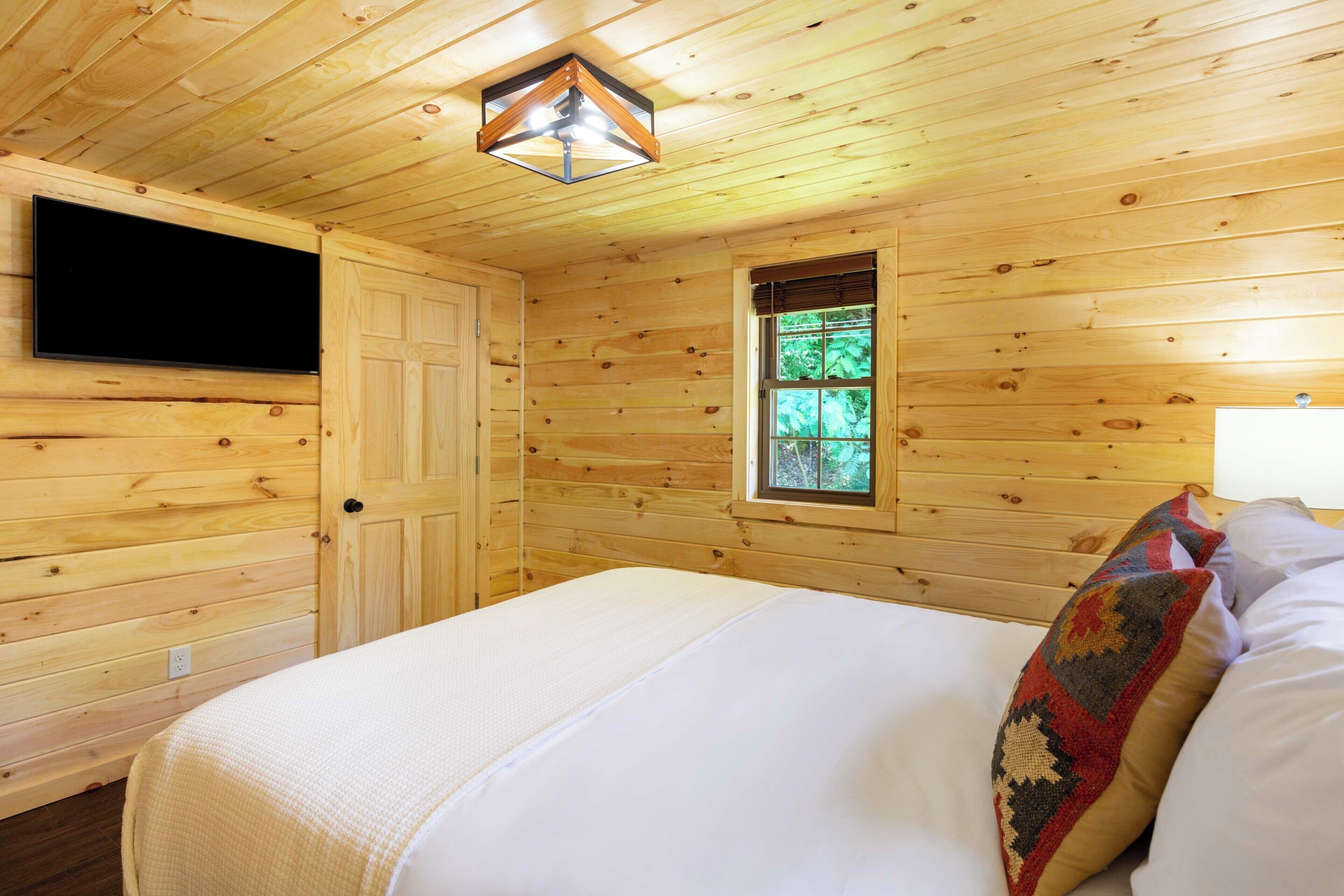 A wooden cabin room with a bed, a TV mounted on the wall, and a window with a view of greenery.