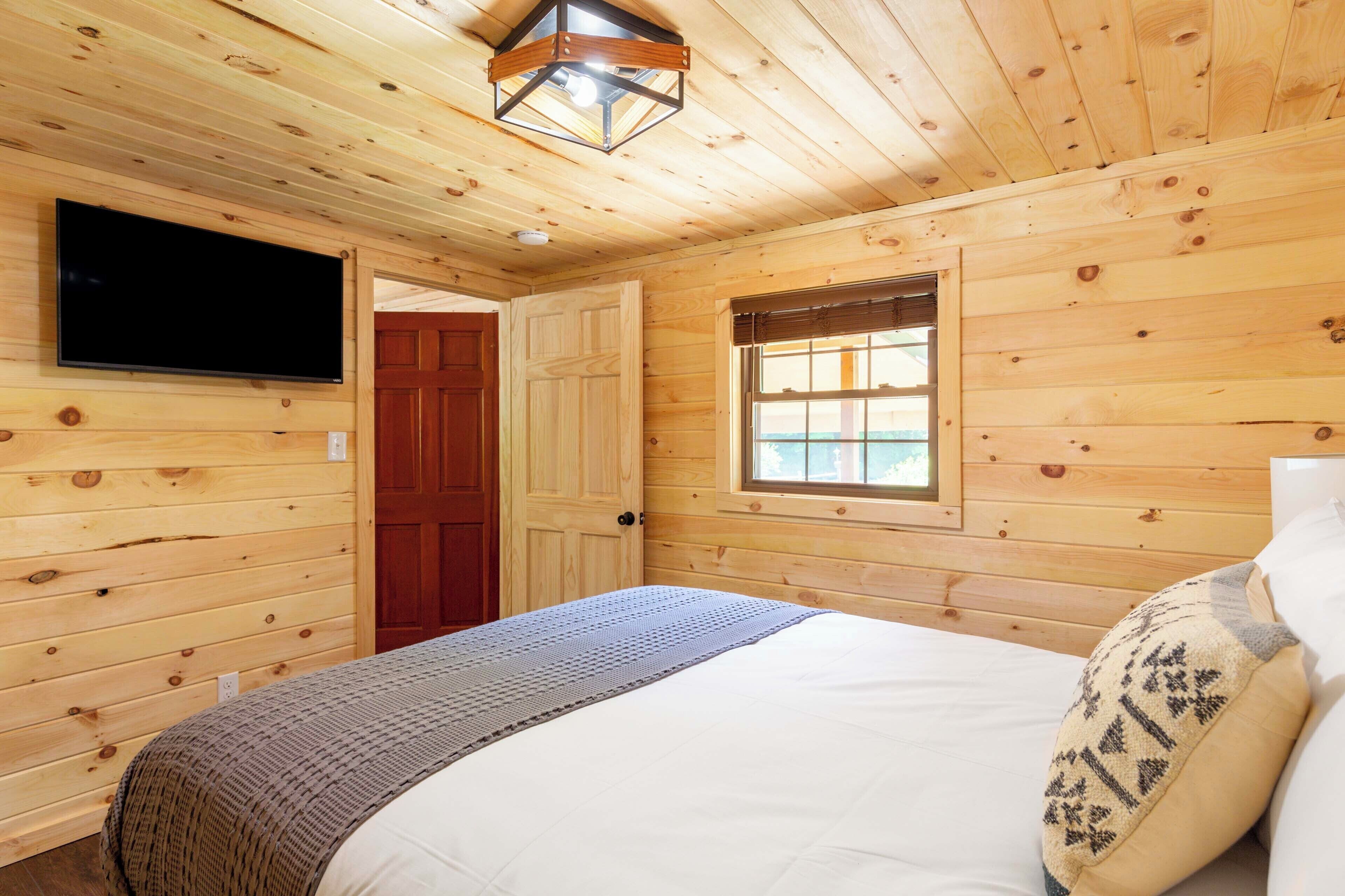 A wooden cabin room with a bed, a television mounted on the wall, and a window.