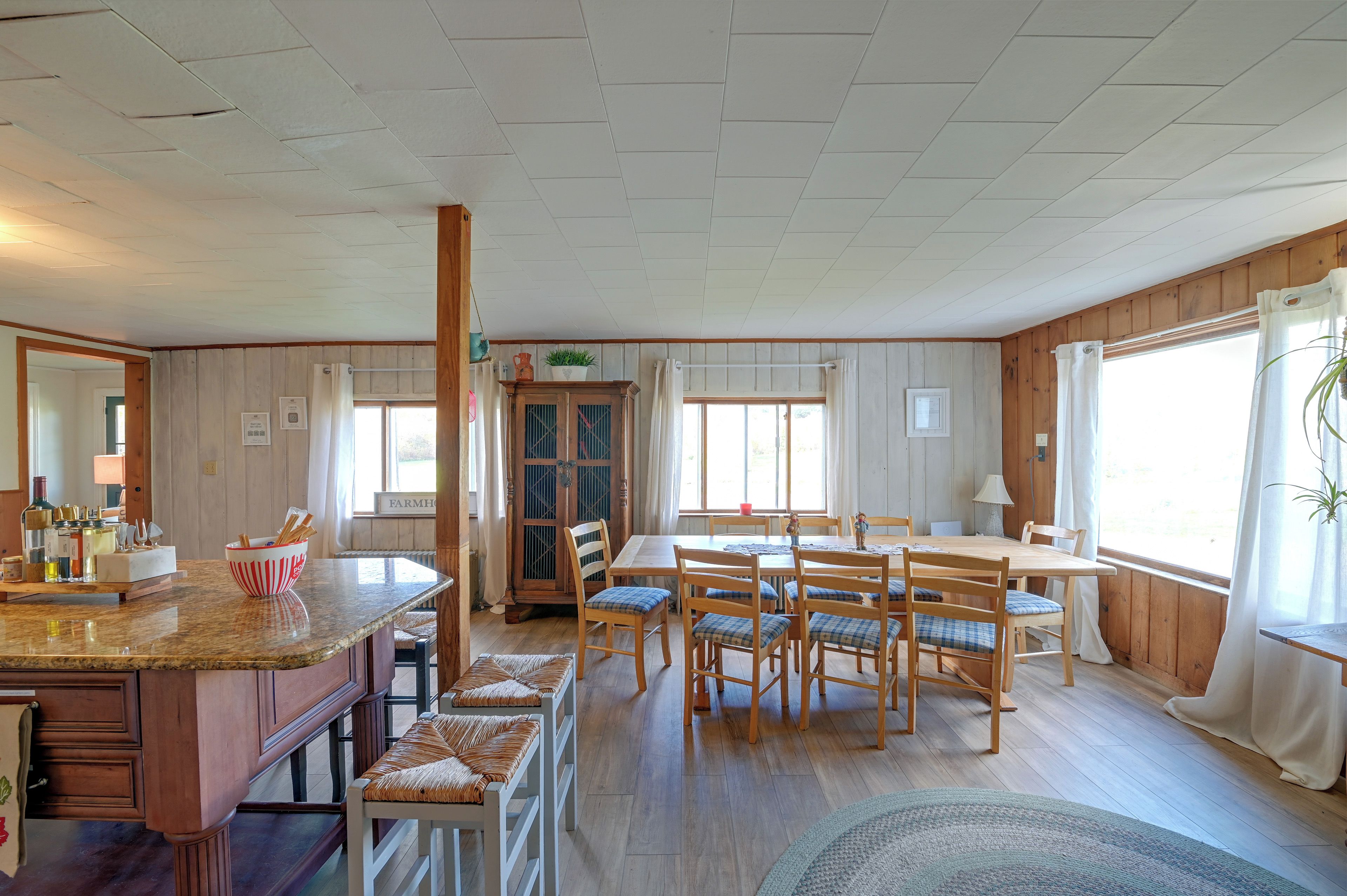 A dining area with a wooden table and chairs, a kitchen counter with a bowl, and a window with curtains.