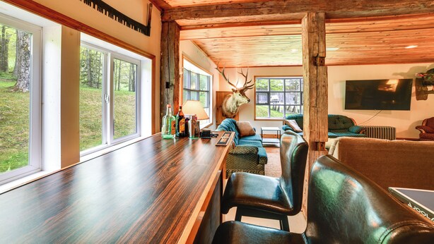 A spacious living area with a wooden ceiling, large windows, a bar with bottles, and a mounted deer head.