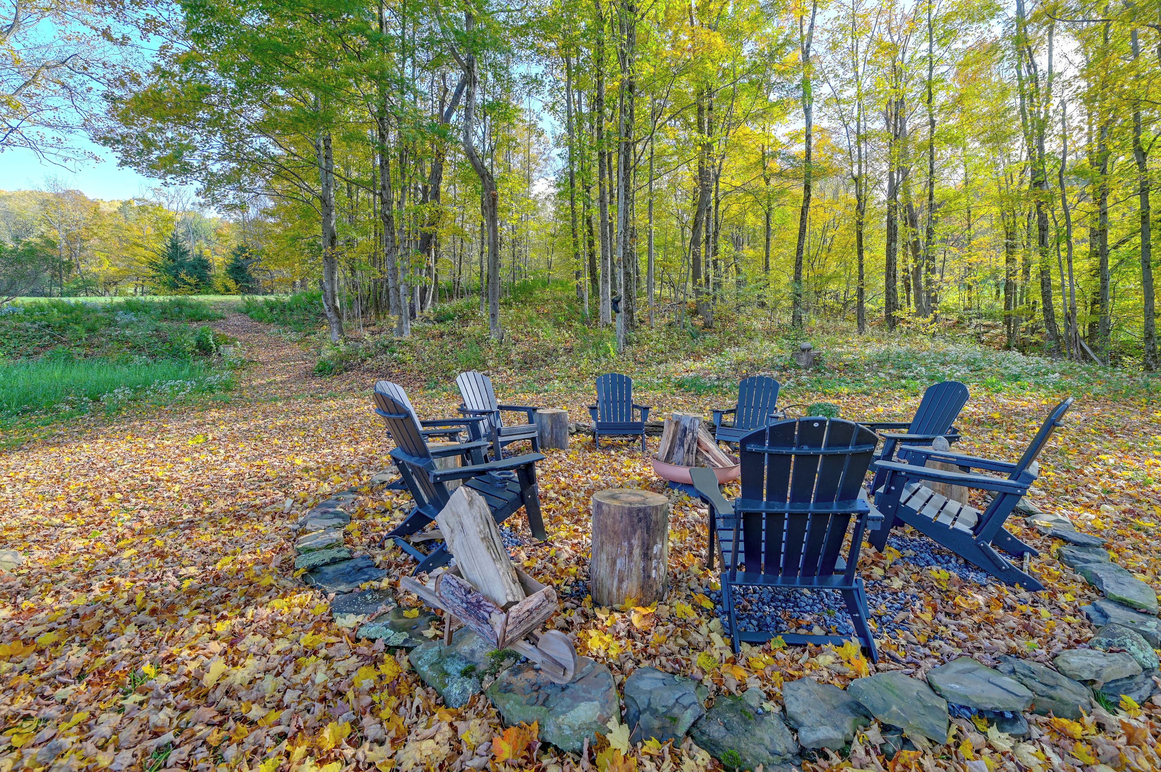 A campsite with four Adirondack chairs arranged around a central fire pit, surrounded by a stone circle and a forest backdrop.