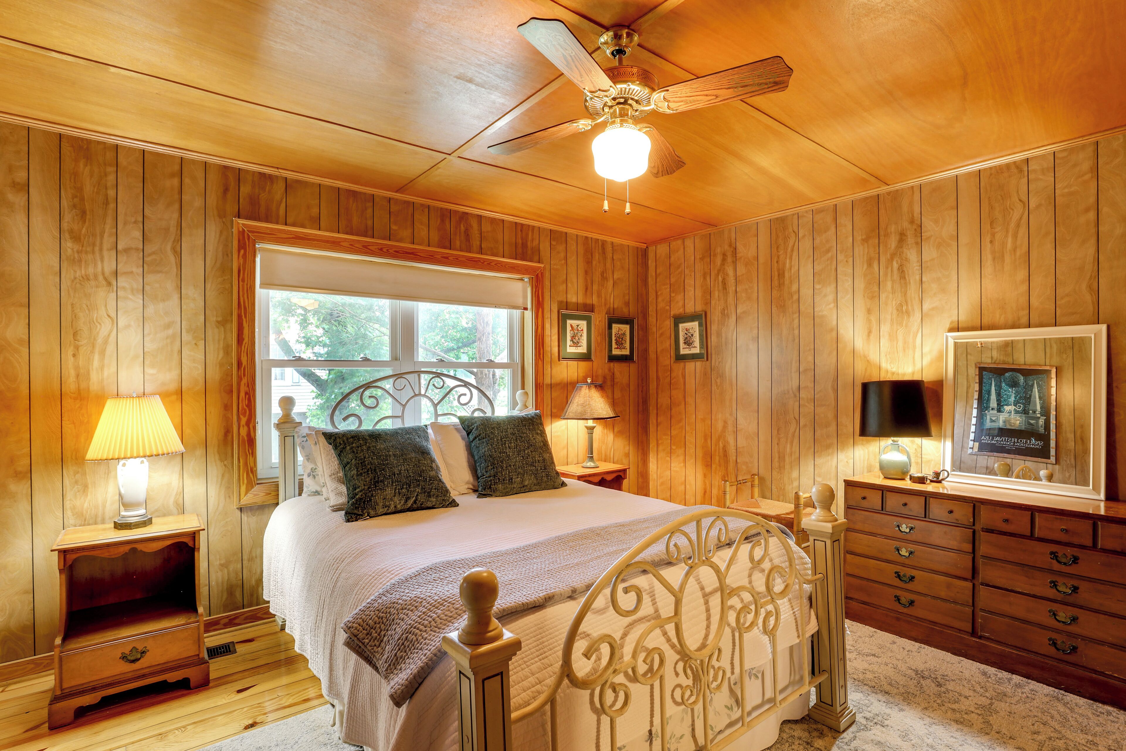 A bedroom with a wooden paneled wall, a bed with a metal headboard, two bedside tables with lamps, a dresser, and a ceiling fan.