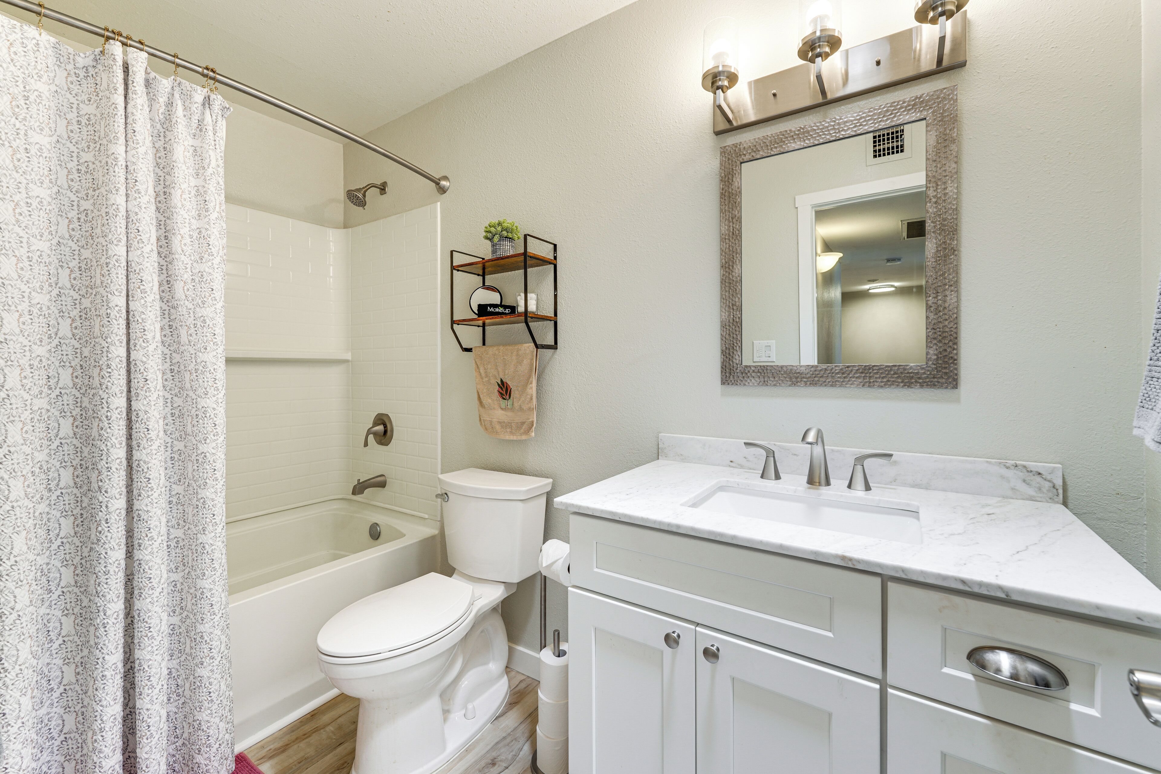 Une salle de bain comprenant des toilettes, une baignoire et un lavabo. On y trouve un rideau de douche, une étagère avec des articles de toilette et un miroir au-dessus du lavabo.